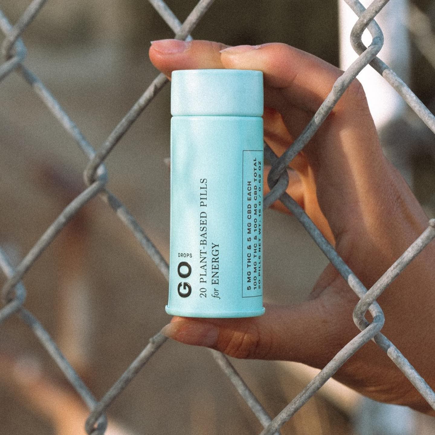 Hand holding a light blue 1906 Go Drops pill tube on the far side of a chain link fence