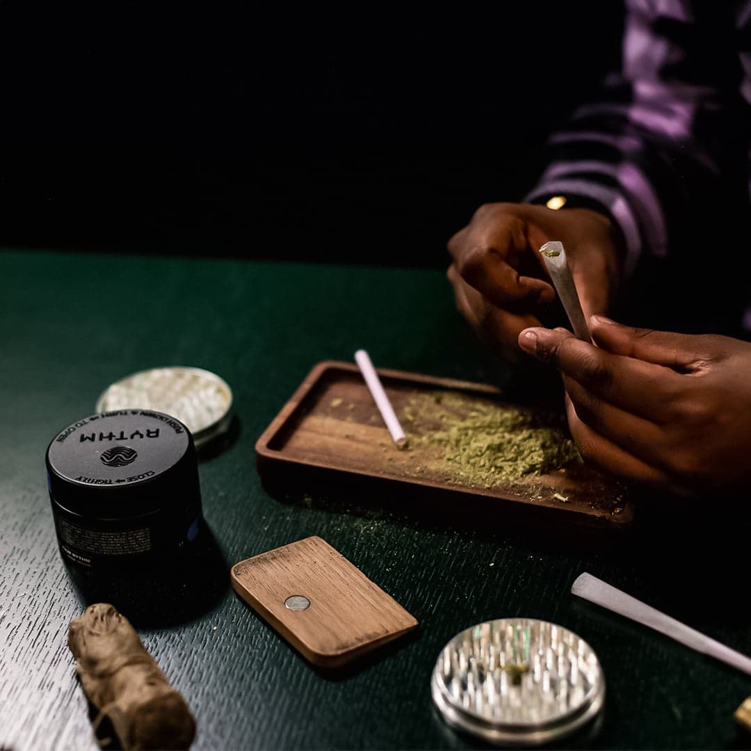 Hands holding a pre-roll over wooden tray with ground cannabis and metal grinder